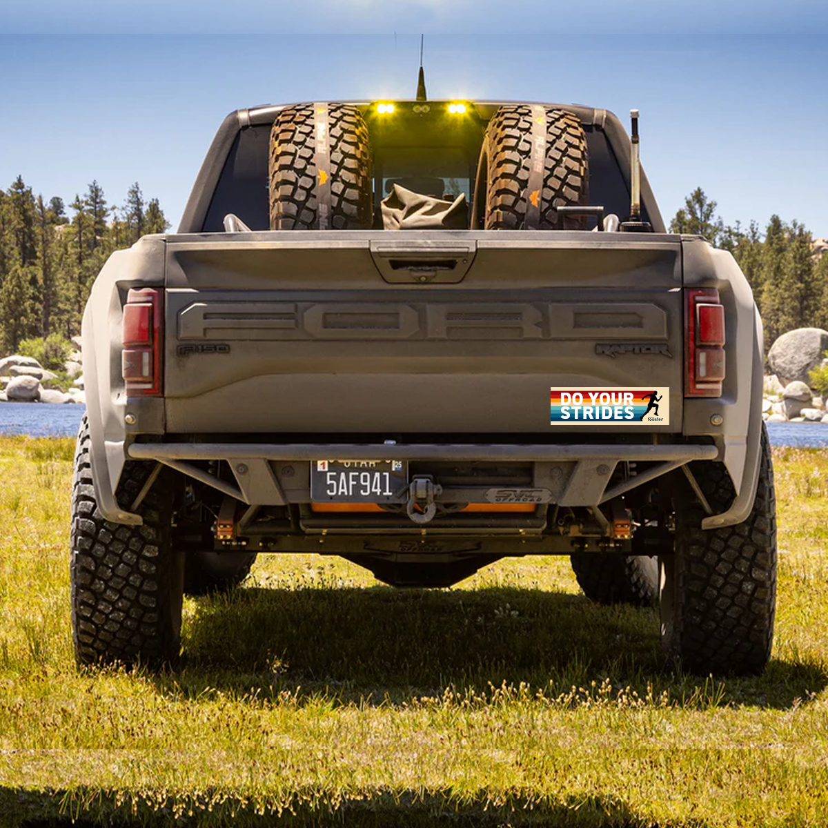Back view of a pickup truck with tires in the bed, parked in a grassy area with trees and water in the background.